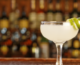 Daiquiri in a glass with lime twist on side sits on bar with blurred out background of liquor bottles behind