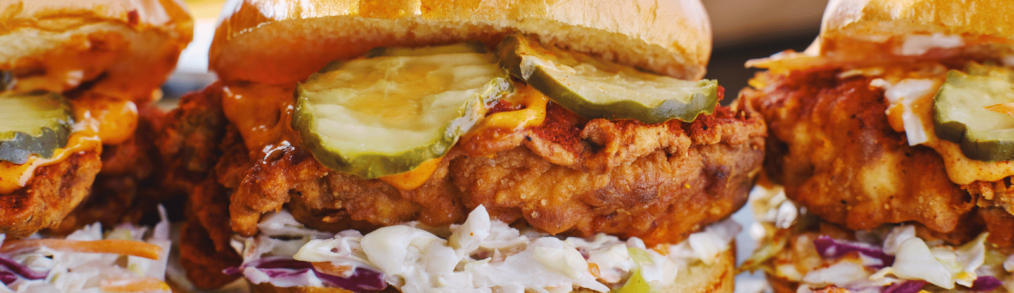 Three fried chicken pieces on buns. Each piece of chicken is sitting on a bed of coleslaw and topped with red sauce and pickles.