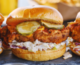 Three fried chicken pieces on buns. Each piece of chicken is sitting on a bed of coleslaw and topped with red sauce and pickles.