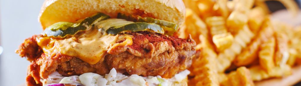 Chicken sandwich with pickles, coleslaw and fixings on wooden board with crinkle French fries on board behind sandwich