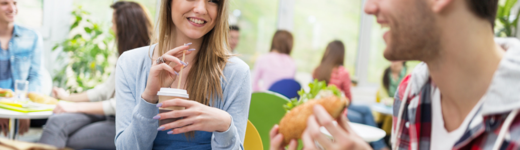 College aged woman with shoulder length hair, light blue sweater and dark blue shirt drinking out of straw in takeout coffee cup. Man with flannel hooded sweatshirt holding hoagie sandwich sitting next to her at table .