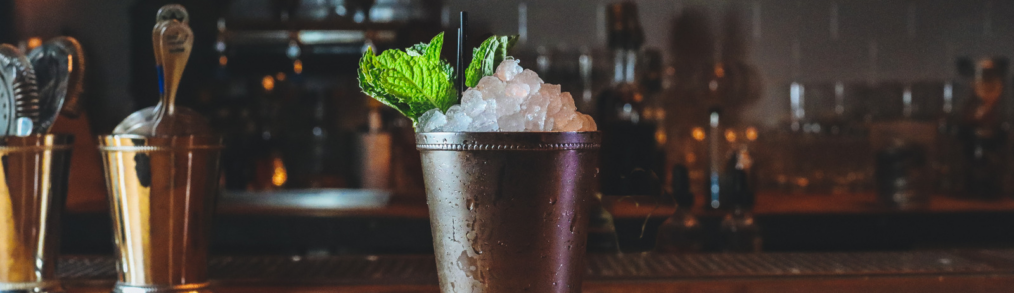 Silver cup on brown bar top. Filled to top with ice, filled with mint julep and sprig of mint on left corner of cup. In background is cocktail shaker and shelfs with liquor bottles lined on them.