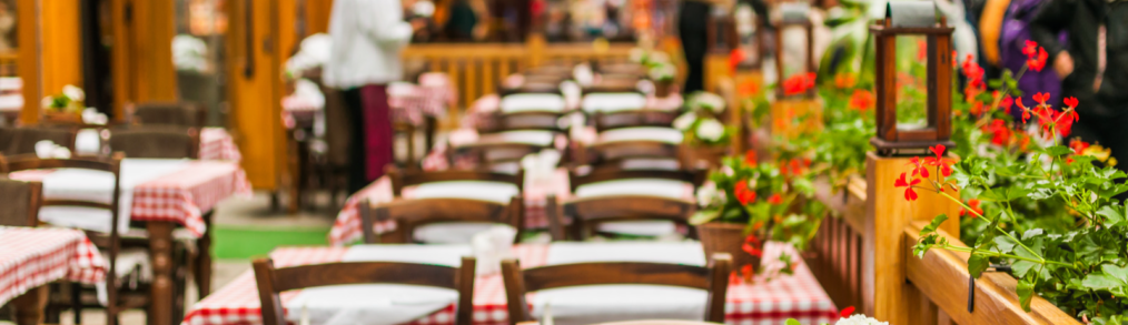 View of brown tables and chairs on outside patio. Waiter in white shirt and red waist apron is in background setting tables while pedestrians walk by on other side of wood railing.