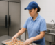 K-12 nutrition worker in school cafeteria wearing light blue shirt, dark blue baseball cap and gloves puts mini pizzas on tray. To worker's right is an industrial refrigerator and behind the worker is a silver sink.