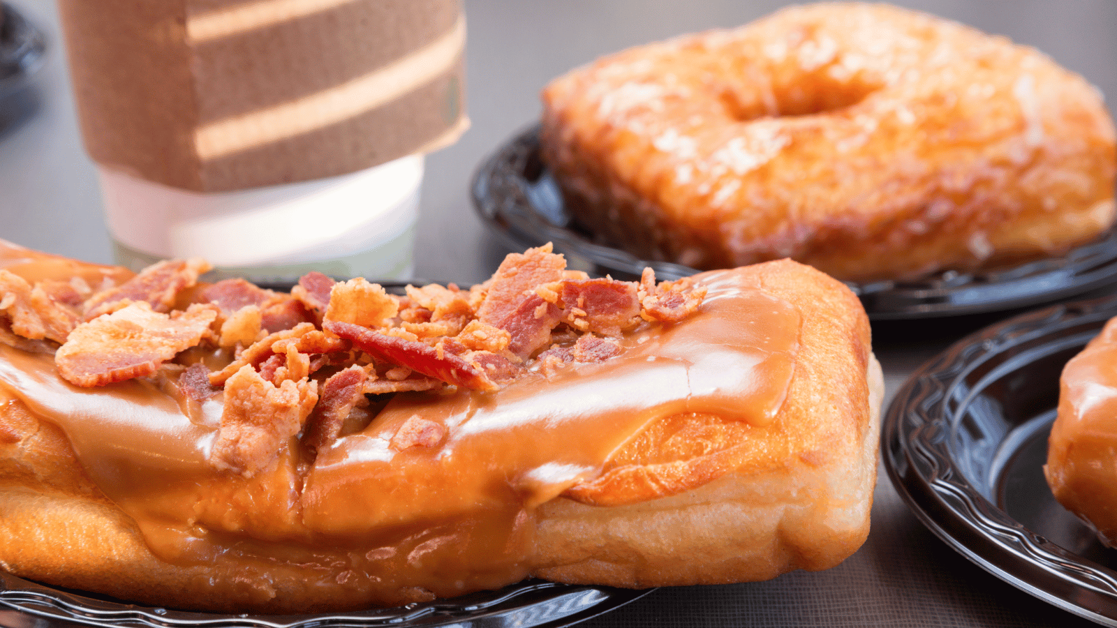 Two donuts, one a round one with a hole and the other a cruller with maple sauce and bacon bits on top. In the background is the bottom of a disposable coffee cup.