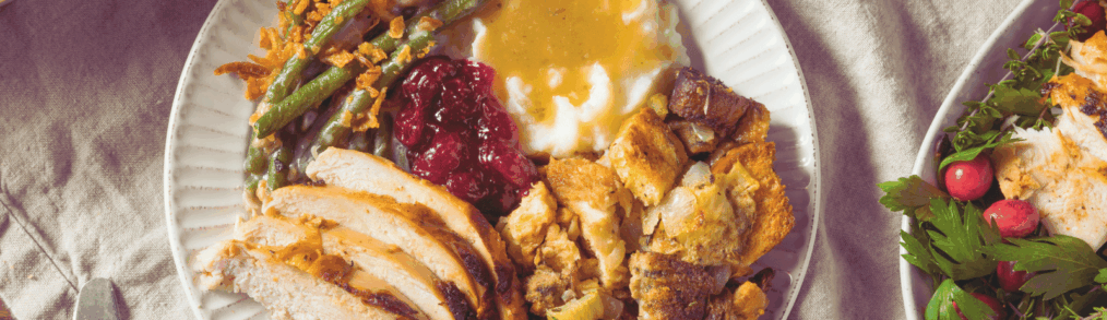 Traditional Thanksgiving foods of sliced turkey, mashed potato with gravy on top, stuffing are on a white plate. Around the plate are bowls of other side dishes.