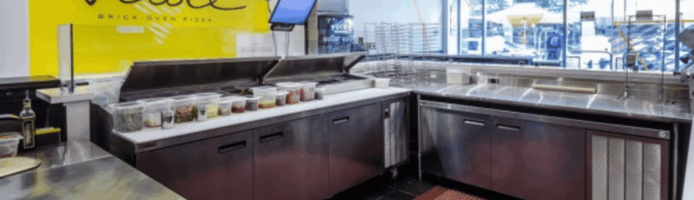 A view into the back area of Veloce pizza showing a stainless steel equipment setup including countertops and refrigeration. Red non-slip mats are on the floor.