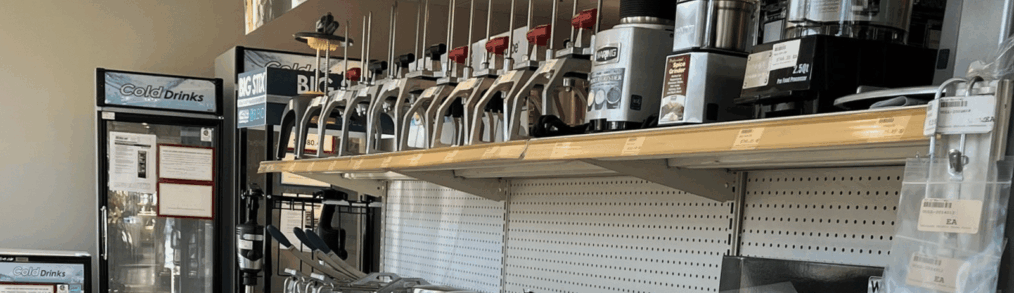 Commercial kitchen equipment dealer showroom displaying countertop appliances, including high-volume blenders, food processors, and prep equipment arranged on retail shelving, with glass-door refrigeration units and merchandising signage in a foodservice supply environment.
