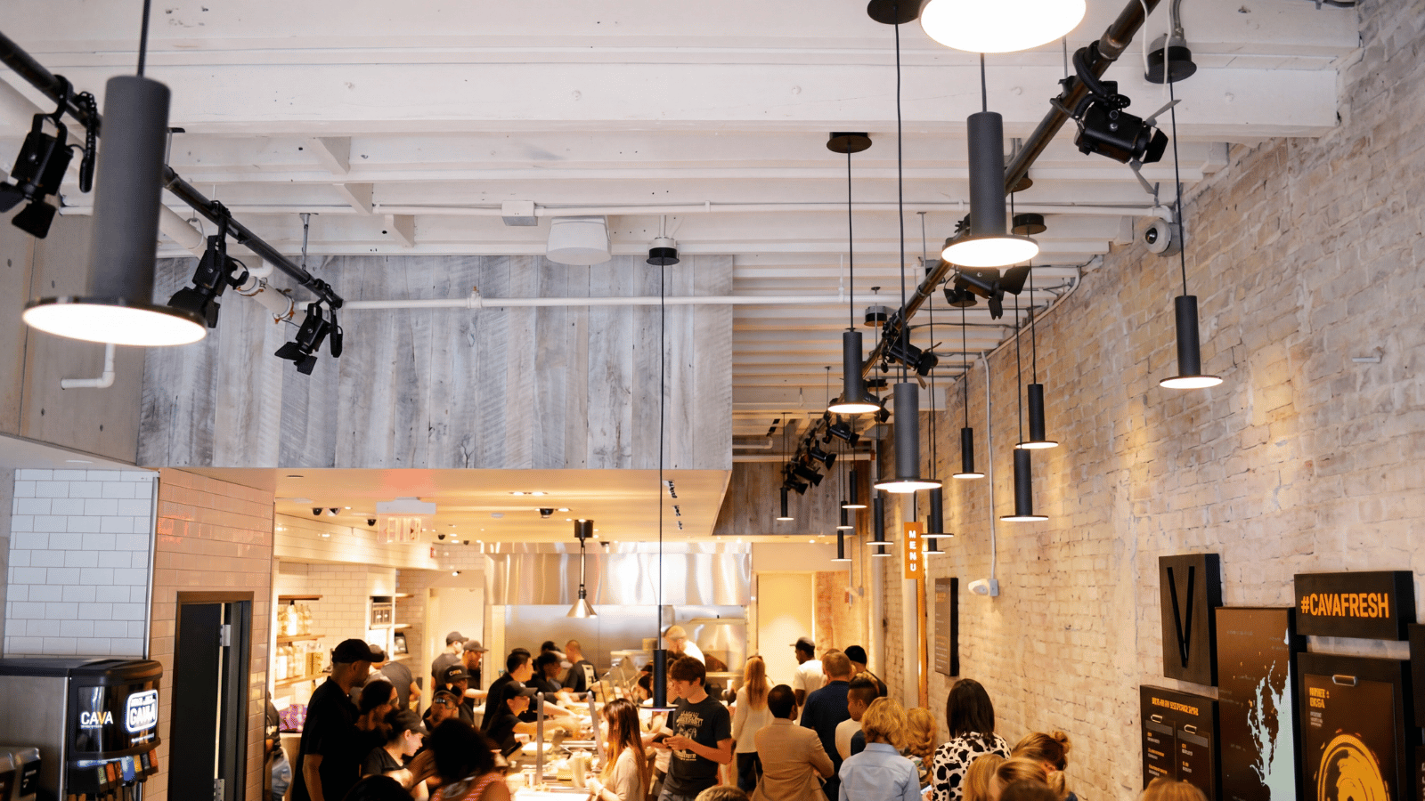 Fast casual restaurant interior with an open commercial kitchen layout, staff actively preparing orders during peak service, customers lined up at the counter, and modern overhead lighting illuminating a high-traffic dining and service area with exposed brick walls.